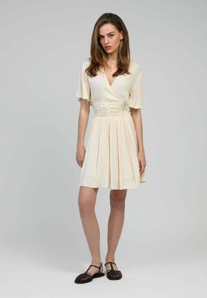 Robe couleur crème avec un décolleté en V et de courtes manches volantées, présentant des détails de taille froncée et une jupe évasée ; associée à des sandales noires.