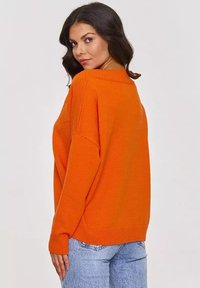 Pomarańczowy sweter dziany o luźnym kroju, szerokim dekolcie i prążkowanych mankietach. Zestawiony z jasnoniebieskimi dżinsami, które mają oversize'owy fason.