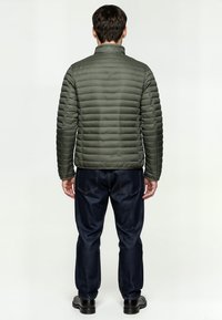 Donkergroene gewatteerde jas met een staande kraag, voorzien van horizontale stiksels en een gladde textuur, gecombineerd met donkerblauwe spijkerbroek en zwarte schoenen.