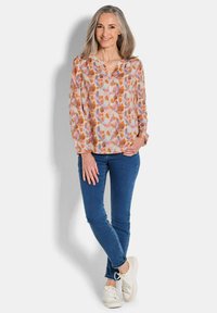 Langärmlige Bluse mit einem rosa und orangefarbenen abstrakten floralen Muster, kombiniert mit blauen engen Jeans und weißen Turnschuhen.