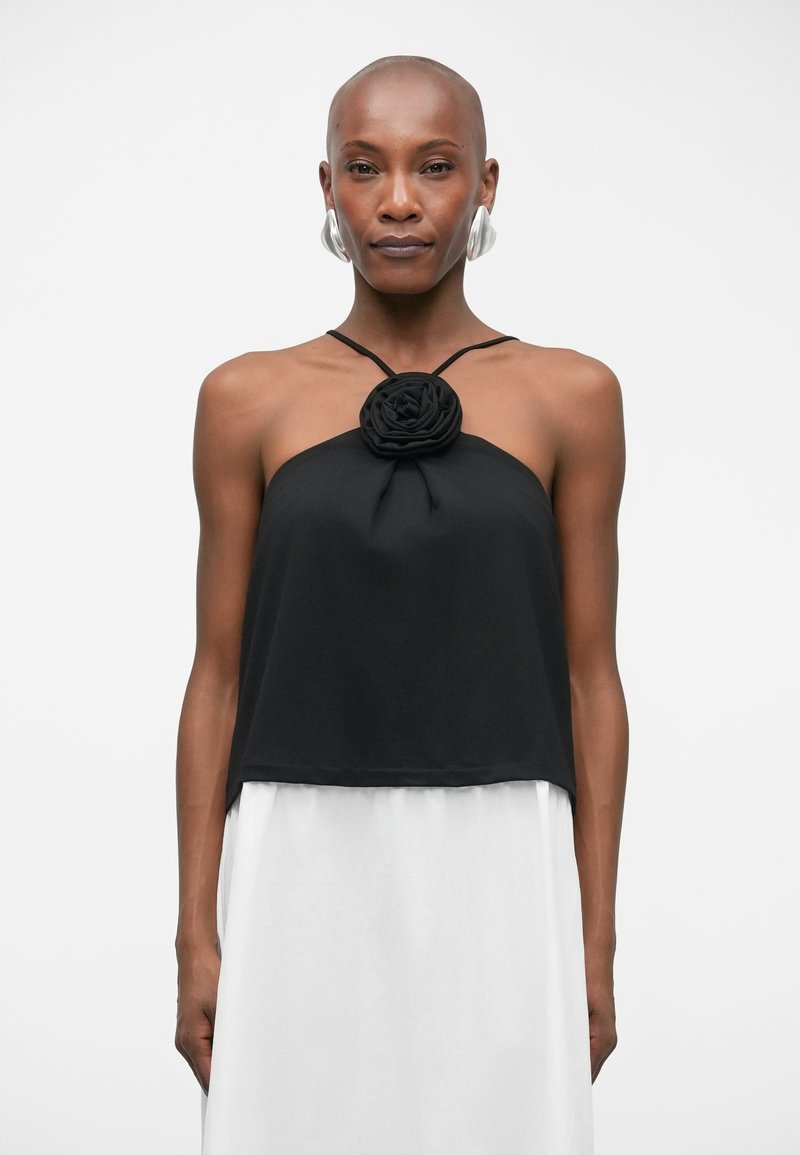 Haut noir à encolure halter avec un détail en forme de rose en tissu au centre, fabriqué dans un matériau doux. Associe à une jupe blanche ; texture globale lisse.