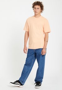 Kortærmet ferskenfarvet t-shirt kombineret med lyseblå løst siddende jeans og sorte sneakers med hvide såler, stående foran en ensfarvet baggrund.