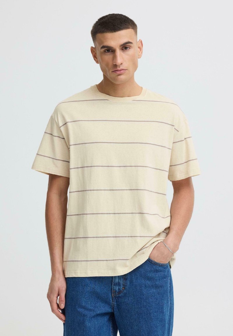 Jeune homme aux cheveux courts portant un t-shirt beige à fines rayures horizontales et un jean bleu, debout avec une main dans la poche.