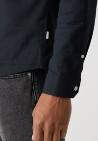 Main d’homme sur le côté portant une chemise noire à manches longues avec poignets boutonnés et un jean en denim gris foncé avec rivets métalliques.