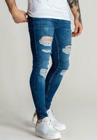 Vaqueros ajustados de hombre en azul con detalles rasgados, que presentan varias zonas desgastadas. Combinados con zapatillas blancas. Diseño ceñido, material de mezclilla.