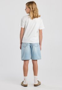 Camiseta blanca de algodón, pantalones cortos de mezclilla azul claro, ajuste casual. Acentuada con calcetines blancos y zapatos marrones sin cordones. Diseño sencillo, sin desorden.