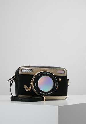 Borsa nera e dorata progettata come una macchina fotografica vintage con obiettivo rotondo con cerniera e tracolla regolabile, posizionata su una superficie bianca.