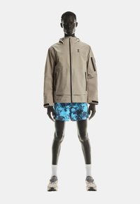 Beige waterdichte jas met een capuchon, met ritszakken, gecombineerd met blauwe gemarmerde shorts en beige sneakers, staand op een effen achtergrond.