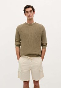Pull en maille vert olive avec texture côtelée, associé à un short cargo beige avec poches latérales et ceinture élastique.
