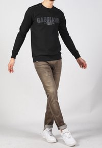 Sudadera negra con texto gris y puños acanalados, combinada con pantalones marrón claro y zapatillas blancas, de pie en un fondo neutro.