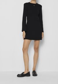 Černé dlouhé šaty s přiléhavým střihem, texturovaná tkanina a malý zlatý akcent na výstřihu, doplněné černými plochými botami.