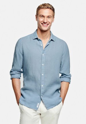 Homme souriant portant une chemise bleu clair boutonnée aux manches retroussées et un pantalon blanc, debout les mains dans les poches sur fond blanc.