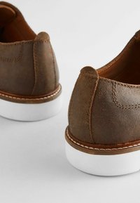 Brązowe zamszowe buty na białej gumowej podeszwie, z przeszyciem wokół podeszwy i miękką, teksturowaną cholewką. Bez sznurowadeł.