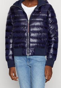 Granatowa kurtka puchowa o błyszczącym wykończeniu, z prążkowanymi mankietami i dołem, zamkiem błyskawicznym oraz kapturem. Charakteryzuje się poziomym pikowaniem.