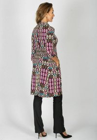 Kobieta stojąca w wzorzystej tunice z długim rękawem w różowe, niebieskie i brązowe romby, czarnych spodniach i czarnych butach na wysokim obcasie.