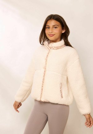 Jeune femme portant une veste en polaire couleur crème avec bordure imprimé léopard et des leggings marron clair, debout devant un fond uni.