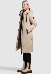 Beige pufferjakke med hætte, chevron-syninger, frontlynlås og sorte detaljer. Båret over sort tøj og parret med beige og sorte støvler.