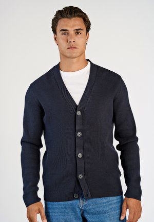 Cardigan a coste di colore blu navy con scollatura a V profonda, chiusura con bottoni e maniche lunghe. Indossato sopra una t-shirt bianca e abbinato a jeans blu.