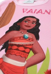 T-shirt rose présentant un personnage aux cheveux foncés et ondulés, portant un haut à motifs rouges et blancs, orné d'un collier bleu. Texte "VAIANI" au-dessus.