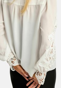 Biela blúzka z priesvitnej látky s prepracovanými čipkovými ozdobami na zvonových rukávoch. Na chrbte je nariasený detail pre zvýraznenie textúry.