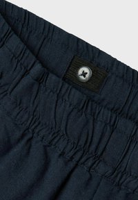 Ceinture en tissu bleu marine avec une texture froncée, dotée d'un bouton argenté et d'un bandeau élastique noir pour un ajustement réglable.