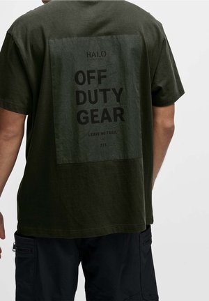 Homme portant un t-shirt vert olive avec une grande tache sombre dans le dos portant l'inscription «OFF DUTY GEAR» et un short cargo noir, debout devant un fond uni.