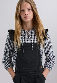 Salopette nera con tasca frontale, indossata su una blusa a scacchi bianco e nero con spalle arricciate e collo a fiocco.