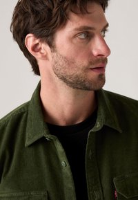 Green corduroy shirt featuring a button-up front, two chest pockets, and a collar. Worn over a black t-shirt with subtle Levi's branding.