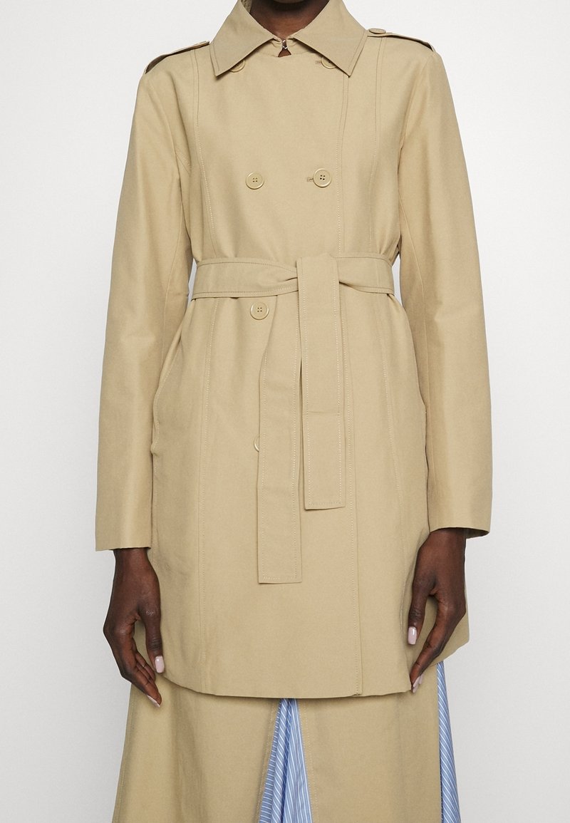 Personne portant un trench-coat beige à double boutonnage avec une ceinture à la taille et des manches longues, debout devant un fond uni.