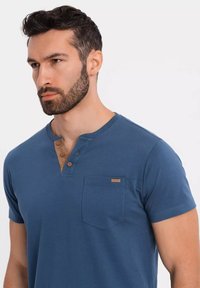 Camisa de algodón azul de manga corta con cuello henley, tres botones marrones y un bolsillo en el pecho que presenta una pequeña etiqueta de logo.