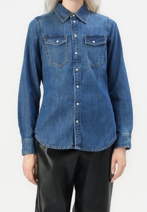 Femme portant une chemise en jean bleue à boutons avec manches longues et deux poches poitrine, associée à un pantalon noir.