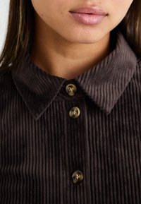 Bruine corduroy overhemd met een kraag, voorzien van verticale ribbels en vier ronde, donkere knopen aan de voorkant.