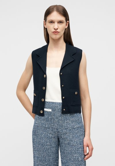 Γυναίκα που φοράει αμάνικο ναυτικό γιλέκο με κουμπιά πάνω από λευκό τοπ, συνδυασμένο με μπλε υφασμάτινο παντελόνι ψηλής μέσης, στέκεται με το ένα χέρι πίσω από την πλάτη.