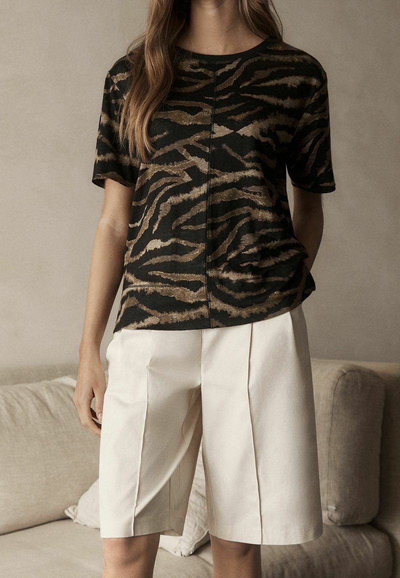 Femme portant une chemise à manches courtes rayée tigre noir et marron avec un short plissé crème longueur genou à l'intérieur.