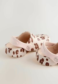 Sabots à imprimé léopard en rose clair et blanc, avec une bride au dos et des semelles texturées, placés sur un fond neutre.