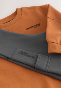 Sweatshirts orange et gris, tissu doux, encolure ronde, poignets et ourlet côtelés. Étiquette de marque visible en noir sur une étiquette grise.