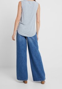 Szary bezrękawnik z zaokrąglonym dołem w zestawieniu z luźnymi, wysokimi niebieskimi dżinsami typu wide-leg. Sandały na drewnianych blokach.