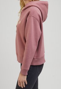 Sweat à capuche rose avec une coupe décontractée, doté d'une capuche à cordon de serrage et de poignets côtelés. Texture douce et design minimaliste sans motifs visibles.