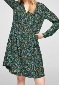 Blommönstrad klänning med svart bas, prydd med gröna, gula och vita blommor. Långa ärmar, V-ringning och en flödande A-linjeformad silhuett.