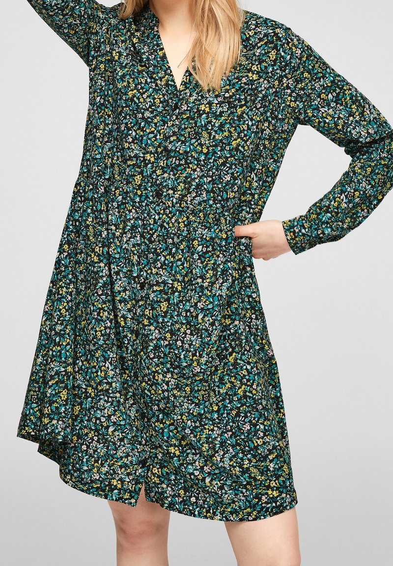 Blommönstrad klänning med svart bas, prydd med gröna, gula och vita blommor. Långa ärmar, V-ringning och en flödande A-linjeformad silhuett.