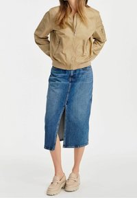 Chaqueta bomber marrón con puños acanalados, combinada con una falda de mezclilla azul con una abertura frontal. Unos zapatos tipo slip-on en beige claro completan el look.