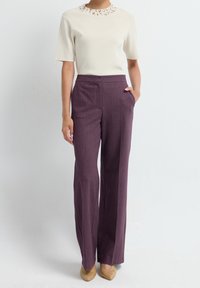Femme portant un haut crème à manches courtes avec un col décoratif, un pantalon violet taille haute à jambes larges, et des chaussures beiges à bout pointu.