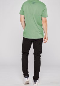 Zelené bavlněné tričko s kulatým výstřihem a krátkými rukávy, v kombinaci s černými džínovými kalhotami a bílými teniskami s námořnickými akcenty.