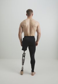 Man med korta svarta atletiska shorts och ett synligt protesben av metall och plast; står barfota, sedd bakifrån.