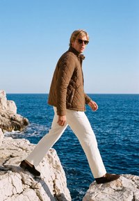 Uomo con giacca trapuntata marrone, pantaloni bianchi e occhiali da sole che cammina tra scogliere rocciose vicino all'oceano blu sotto un cielo sereno.