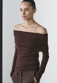 Brown off-the-shoulder top made of soft fabric with long sleeves and draping details, paired with dark trousers. Smooth texture.