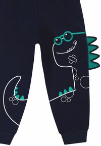Jogging pants navy con grafica di un dinosauro bianco, con accenti turchesi sulle spine e sugli occhiali, e motivi circolari vicino alla coda. Materiale in cotone.