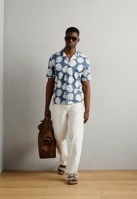 Korte mouwen blauw shirt met witte bladerenpatronen, witte broek, leren bruine tas, bruine sandalen en zonnebril. Minimalistische achtergrond.