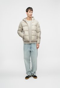 Beige pufferjakke med hætte, der har quiltede sektioner. Båret over en lysegrå top og parret med løstsiddende lys blå jeans og sorte sneakers.