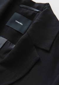 Zwarte op maat gemaakte blazer van een textuurstof, met scherpe revers en een ingenaaide merklabel met "someday" in het wit op een donkere achtergrond.